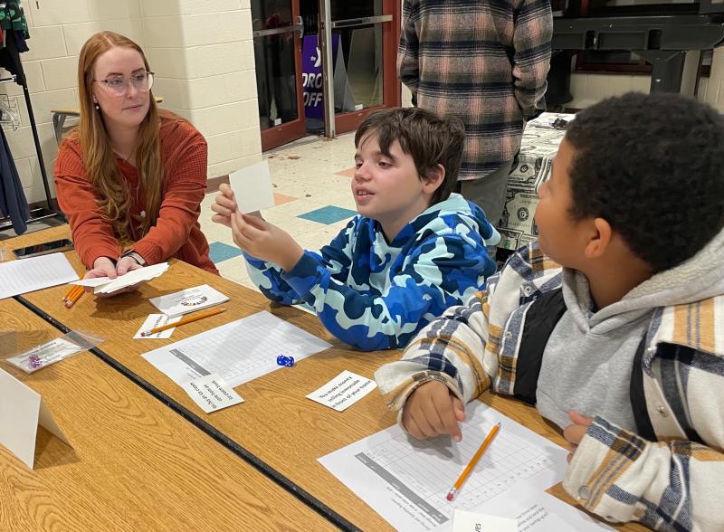 Fourth-grade teacher Marissa LeGates helps Cash Erickson and Issac Norwood draw cards to play the saving-and-spending game.